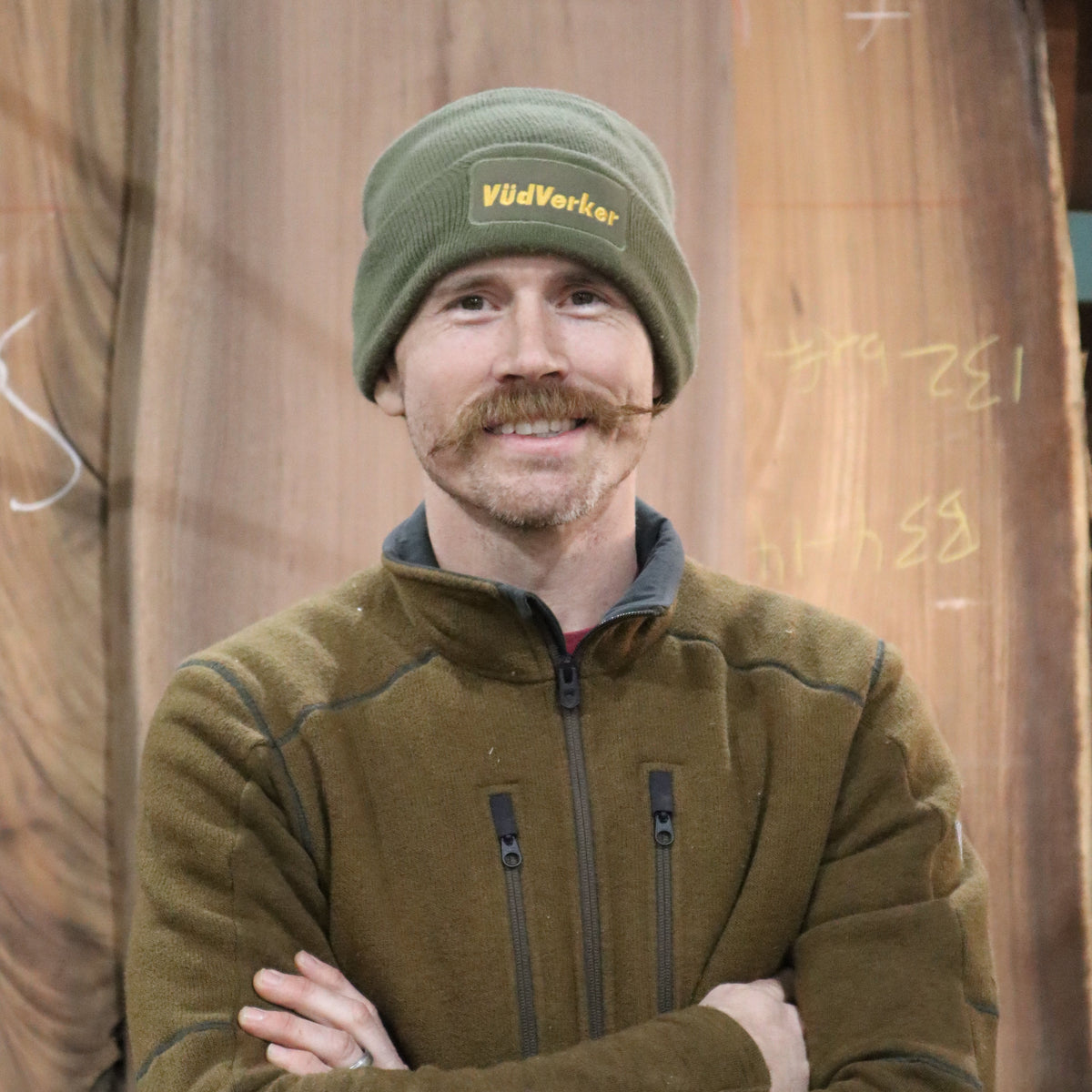 A man wearing an olive green fleece zip-up jacket and a matching green beanie hat with yellow embroidered text, standing with arms crossed in front of a large wood slab background.