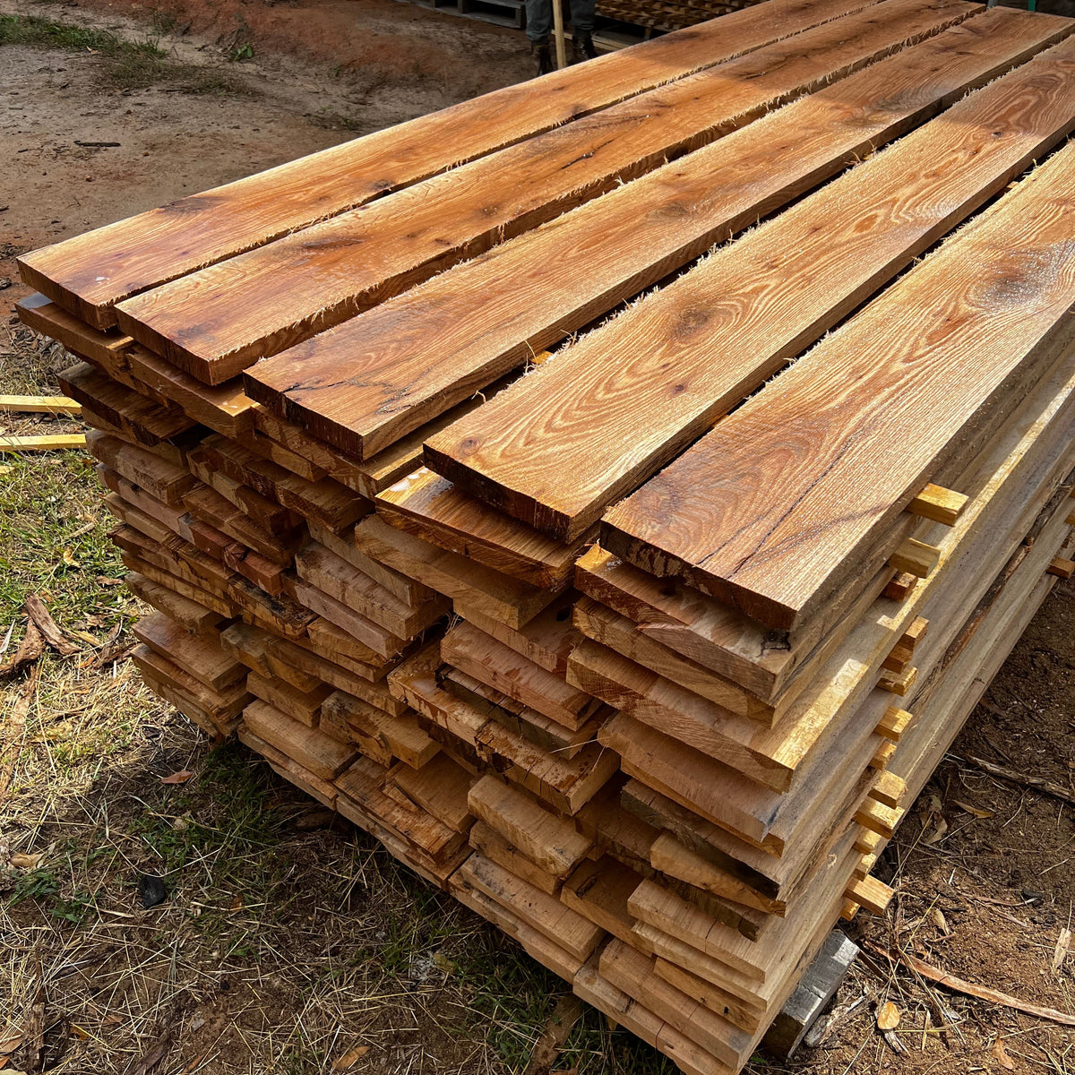A large stack of raw wooden planks with natural grain and live edges, piled outdoors on a dirt and grass ground.